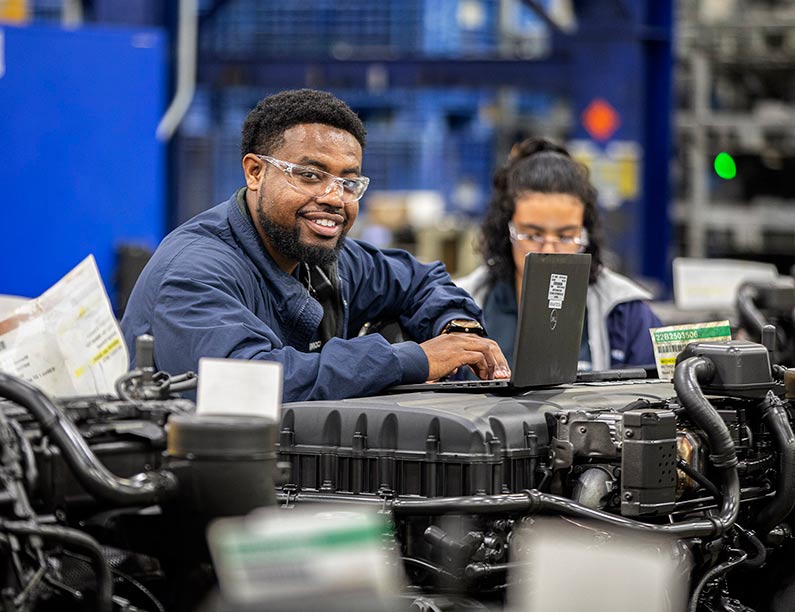 Two workers in a factory with one wearing protective eyewear and using a computer
